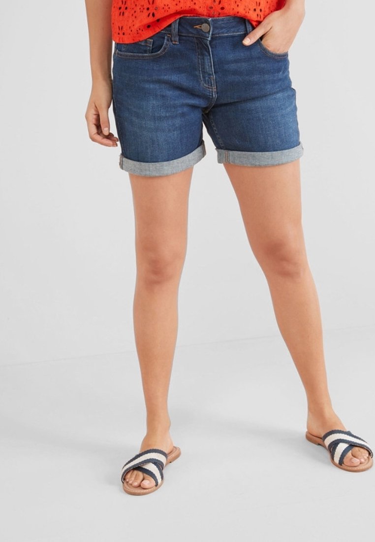 Nainen, joka on pukeutunut käännettäviin tummiin farkkushortseihin, oranssiin puseroon ja raidallisiin liukuvaan sandaaleihin, seisoo käsi yhdessa taskussa.