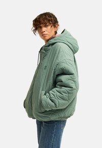 Woman with curly hair wearing oversized green quilted hooded jacket and blue jeans, hands in pockets, looking toward camera.
