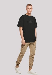 Zwart T-shirt met korte mouwen en een grafische afbeelding van bergen op de borst, gecombineerd met beige cargobroeken en witte sneakers. Casual, relaxte pasvorm.