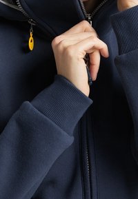 Marinblå zip-tröja med ribbade muddar. Har en hög krage, silverfärgad dragkedja och en gul dragkedjeanordning för enkel stängning. Mjuk textur.