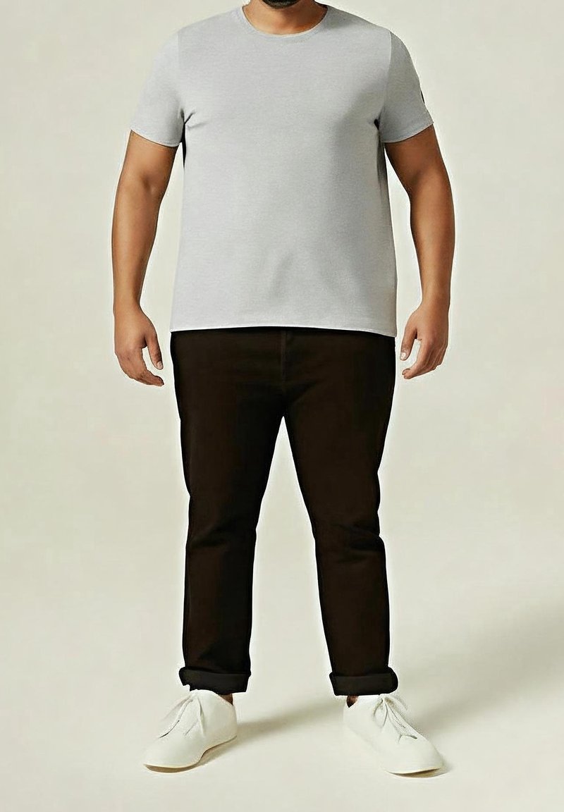 Homme portant un t-shirt gris clair à manches courtes, un pantalon noir retroussé et des chaussures blanches à lacets, debout sur un fond uni.