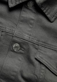 Grå denimtyg med en strukturerad yta, med en knappstängning och en bröstficka. Sömnadsdetaljer är synliga längs sömmarna.