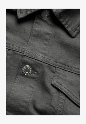 Grauer Denim-Stoff mit einer strukturierten Oberfläche, versehen mit einem Knopfverschluss und einer Brusttasche. Nähte sind entlang der Säume sichtbar.