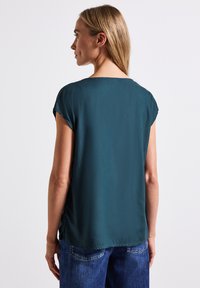 Blusa verde acqua con una texture liscia simile alla seta, maniche a cappuccio e scollatura ampia. Presenta spacco laterale e una vestibilità rilassata. Abbinata a jeans in denim.