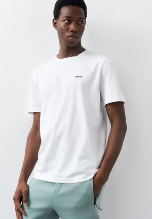 Νεαρός άνδρας που φοράει λευκό μπλουζάκι με την επιγραφή "BOSS" και ανοιχτό πράσινο παντελόνι, στέκεται με το ένα χέρι στην τσέπη, μπροστά σε απλό λευκό φόντο.