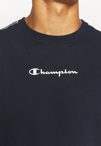 Camiseta de algodón azul marino con cuello redondo y logo "Champion" blanco en el pecho. Incluye detalles de rayas laterales en contraste.