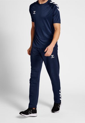 Homme portant une tenue de sport hummel bleu marine avec des détails chevrons blancs sur les épaules et le pantalon, ainsi que des chaussures de sport noires sur un fond uni.
