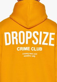 Gul hoodie med hvid tekst. Har "DROPSIZE" øverst, "CRIME CLUB" nedenunder og mindre tekst: "LEGENDS SINCE 2020, kun for medlemmer." Blød stofstruktur.