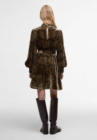 Πράσινο φόρεμα βελούδινο με ανθισμένο μοτίβο, μακριά φουσκωτά μανίκια, άνοιγμα στην πλάτη και στύφωμα στη μέση, συνδυασμένο με καφέ μπότες μέχρι το γόνατο.