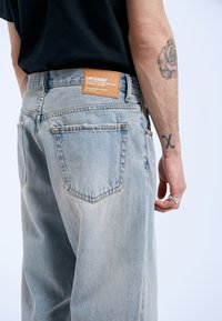 Jeans de mezclilla azul claro con un corte relajado, que cuentan con una etiqueta de marca de cuero marrón y dos bolsillos traseros, sobre un fondo liso.