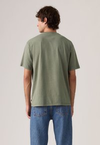 Άνδρας που φοράει ελιάς πράσινο T-shirt και μπλε τζιν, στέκεται με την πλάτη προς την κάμερα μπροστά σε ένα απλό φωτεινό φόντο.