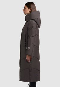Profiel van een vrouw die een lange donkere pufferjas met hoge kraag, capuchon, ritszakken en drukknoopjes langs de zijnaad draagt.