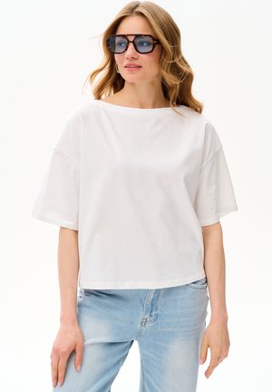 Kobieta z długimi blond włosami, nosząca duże okulary w wzór żółwia, białą luźną koszulkę z krótkim rękawem oraz jasnoniebieskie dżinsy, patrząca w bok.