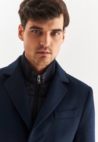 Granatowy płaszcz z wełny z domieszką, z wyłogami w kształcie ostrosłupa i zapinaną na zamek wewnętrzną warstwą, charakteryzujący się eleganckim fasonem i jedną kieszenią na piersi.