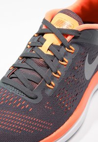 Šedá sportovní obuv s mřížkovou tkaninou, oranžovými akcenty a šedým znakem swoosh. Má šněrovací design a texturovanou podrážku pro lepší přilnavost.