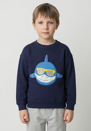 Marineblauwe sweatshirt met een grote, lachende haai met gele zonnebril op de voorkant. Grijze joggingbroek maakt de outfit compleet.