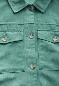 Chemise en velours côtelé teal avec col, deux poches avant et boutons ronds. Le tissu a une texture côtelée. Fils visibles près des boutons.