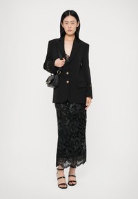 Svart blazer med guldknappar, bärs över en spetsklänning med blommönster och en scallop-kant. Modellen håller en liten svart handväska.
