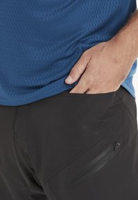 Sininen tekstuurillinen lyhythihainen paita yhdistettynä mustiin housuihin, joissa on vetoketjullinen tasku, sileä kangas ja istuva muoto.