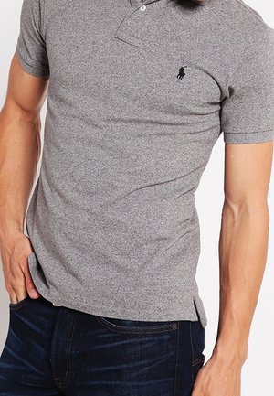Man draagt een nauwsluitend grijs poloshirt met een klein zwart paardenlogo, gecombineerd met donkerblauwe spijkerbroek, hand rustend in de zak.