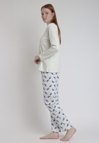 Blusa de manga larga en color crema claro combinada con pantalones de pijama blancos que presentan un patrón repetido de flores y rayas, textura suave y corte recto.