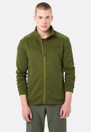 Junger Mann trägt eine olivgrüne Fleecejacke mit Reißverschluss und langen Ärmeln sowie passende grüne Hose, steht vor weißem Hintergrund.