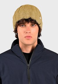 Breipet in bruin met een ribbeltextuur. Draagend door een model met donker haar, gecombineerd met een marineblauwe jack met rits, met een hoge kraag.