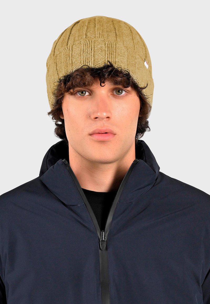 Breipet in bruin met een ribbeltextuur. Draagend door een model met donker haar, gecombineerd met een marineblauwe jack met rits, met een hoge kraag.