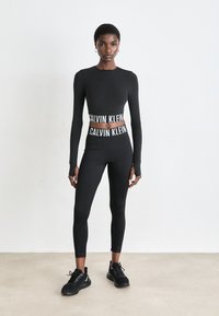 Frau in schwarzer Calvin Klein Activewear mit langen Ärmeln, hoch geschnittenen Leggings und schwarzen Turnschuhen vor einem schlichten Hintergrund.