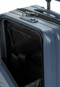 Valigia rigida blu navy con compartimento frontale aperto, dotata di tasche interne, cintura nera e serratura a combinazione TSA integrata nella parte superiore.