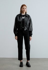 Μαύρο τζάκετ από συνθετικό δέρμα με φερμουάρ μπροστά, φορεμένο πάνω από μαύρο τοπ και μαύρο παντελόνι με ψηλή μέση. Το μοντέλο φοράει μαύρα παπούτσια πλατφόρμας.