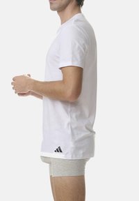 White cotton T-shirt with short sleeves, featuring a black Adidas logo on the left side and a relaxed fit. Grey underwear is visible.