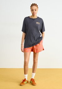 T-shirt oversize gris foncé avec le logo Roxy, associée à un short à motifs rose vif et des baskets orange avec des accents verts. Chaussettes blanches visibles.