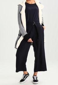 Långa svarta byxor med lös passform, kombinerade med en marinblå topp och en grå stickad cardigan med ett mysigt, vitt fleecefoder och svarta manschetter.