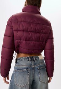 Burgundy cropped pufferjakke med høj krave, featuring horisontale sømme og en snoretræk i kanten, båret over lyseblå jeans.