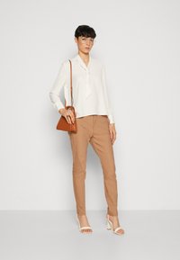 Weißes Blusenshirt mit Knotendetail am Kragen, kombiniert mit hellbraunen, schmal zulaufenden Hosen. Das Model hält eine kleine braune Tasche und trägt weiße Absatzsandalen.