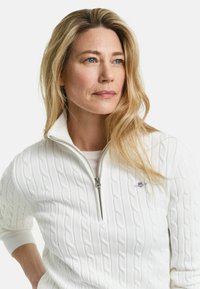 Ξανθιά γυναίκα με μπλε μάτια που φοράει λευκό πουλόβερ με πλέξη κοτσίδα και γιακά με φερμουάρ μέχρι τη μέση, κοιτάζει λίγο προς τα δεξιά σε λευκό φόντο.