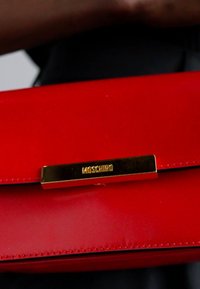 Soehest punane nahkne kott, millel on kuldtooni klamber, millel on graveeritud "Moschino". Sujuv tekstuur ja stiliseeritud ristkülikukujuline kuju.