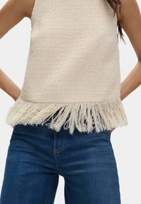 Canotta di colore crema con orlo frangiato. Tessuto intrecciato, design senza maniche, abbinata a jeans blu scuro.