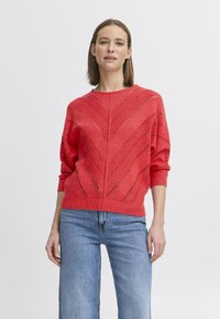 Roter Strickpullover mit Zickzackdesign, der offene Strickdetails sowie gerippte Bündchen und Saum aufweist. Kombiniert mit blauen weiten Jeans.