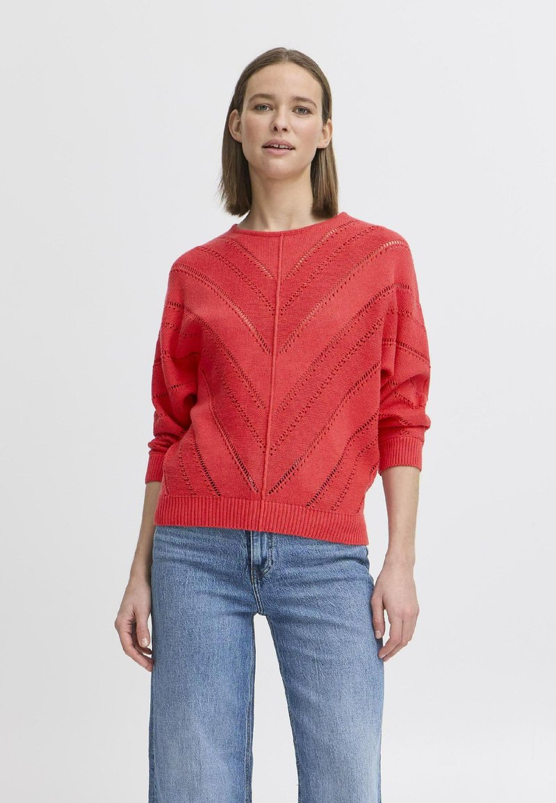Roter Strickpullover mit Zickzackdesign, der offene Strickdetails sowie gerippte Bündchen und Saum aufweist. Kombiniert mit blauen weiten Jeans.