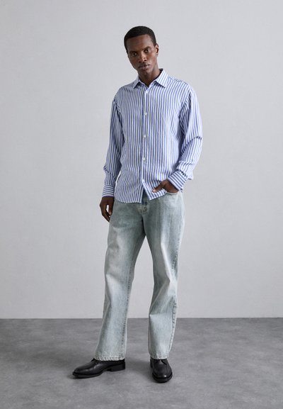 Camisa de botones a rayas azul y blanca con cuello, combinada con jeans de lavado claro y zapatos negros. Tejido suave y corte relajado.
