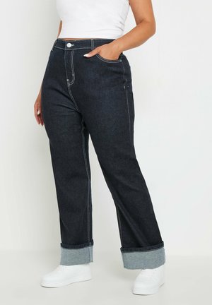 Dunkelblaue Jeans mit geradem Schnitt, ausgestattet mit weißen Nähten und umgeschlagenen Säumen, getragen mit weißen Sneakern.