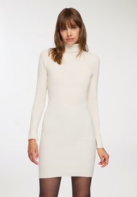 Abito a collo alto a coste crema con maniche lunghe. Silhouette aderente e lunghezza sopra il ginocchio. Texture liscia e design minimal.
