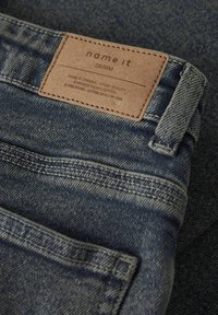 Denimjeans med en brun sydd etikett som sier "name it DENIM" over baklommen og beltestroppen på mørkeblått stoff.