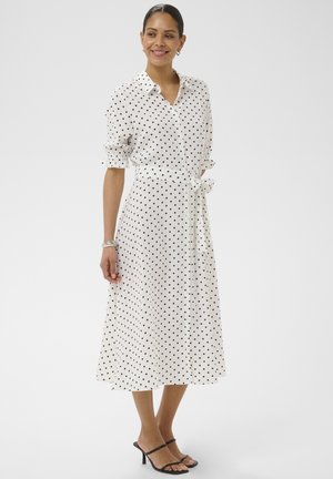 Kobieta uśmiechnięta, ubrana w białą sukienkę midi w czarne groszki, z podwiniętymi rękawami, paskiem w talii i czarnymi sandałami na obcasie na jednolitym tle.