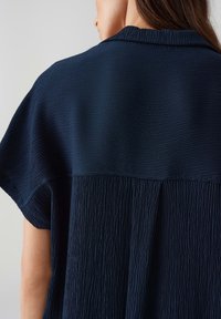 Top blu navy con un tessuto testurizzato che presenta una parte superiore liscia e una parte inferiore plissettata. Maniche corte e un design con colletto morbido.