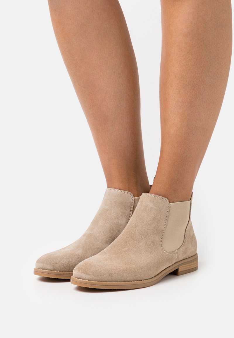 Bottines en daim beige avec panneaux latéraux élastiques portées sur des jambes nues, sur un fond blanc uni.