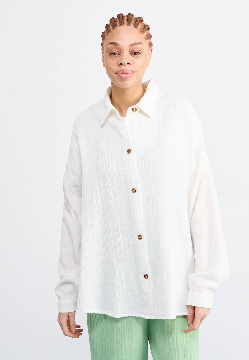 White button-up shirt made of textured fabric, featuring a collar and brown buttons. Worn over green, pleated pants.
