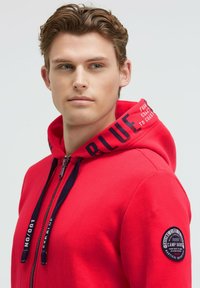 Sudadera roja con cremallera y cordones azul marino, con texto impreso en la capucha. Presenta un parche redondo con la marca en la manga izquierda. Tejido suave.